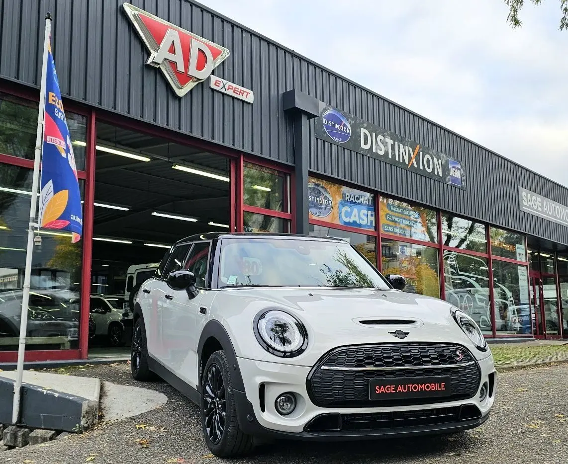 Vue 3/4 avant d'une MINI Cooper S Clubman blanche avec toit noir, garée devant un garage Sage Automobile.