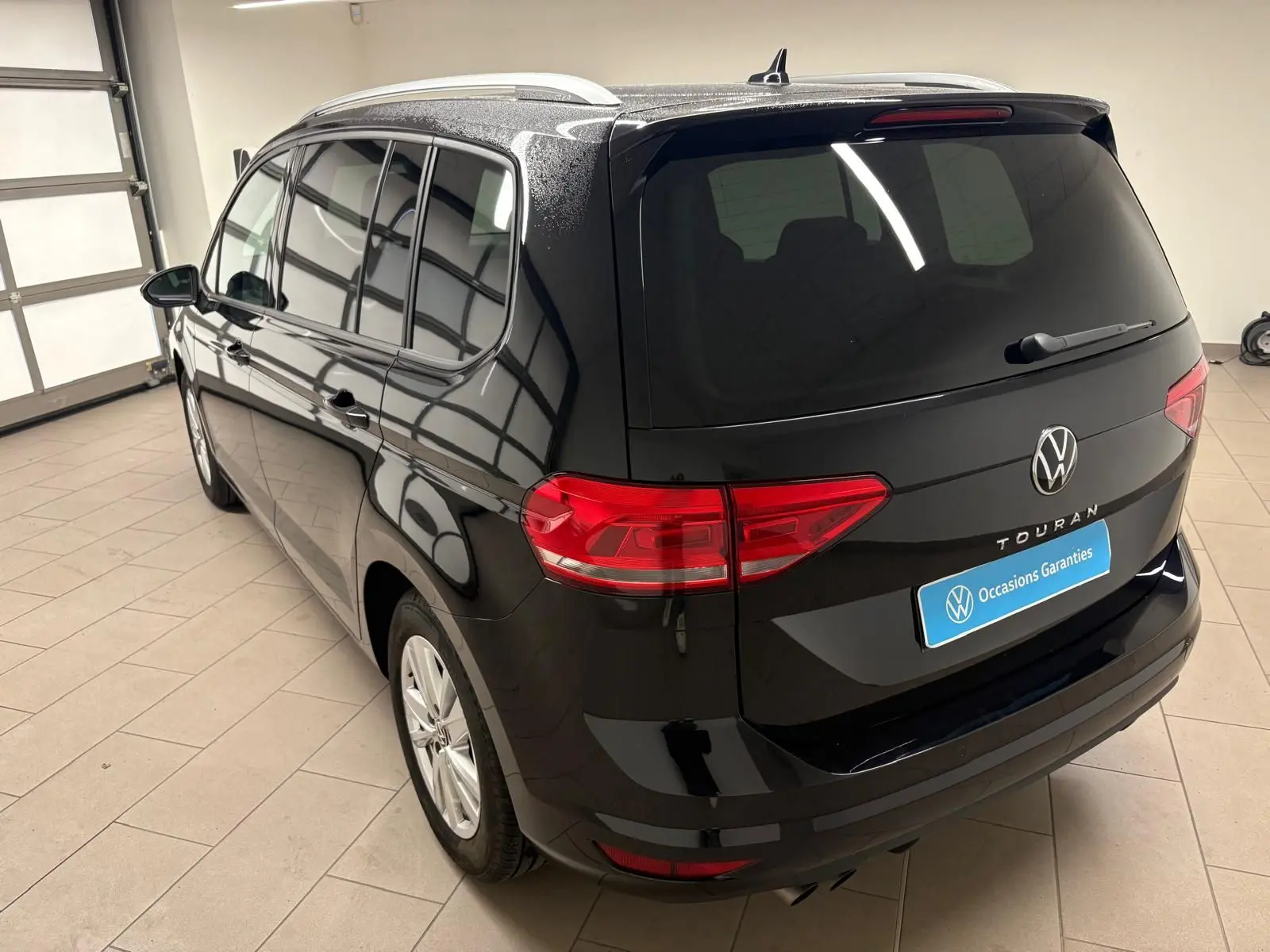 Vue 3/4 arrière gauche d'un Volkswagen Touran noir intense nacré avec hayon électrique et vitres teintées dans un garage.