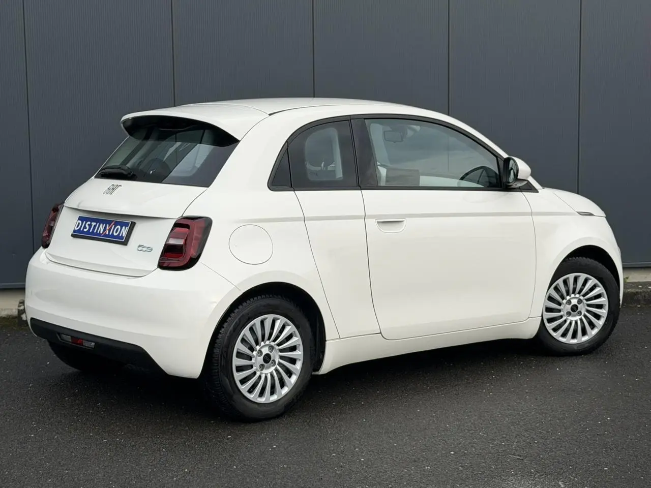 Vue 3/4 arrière droite de la FIAT 500 électrique blanche 3+1 avec jantes alliage et toit noir contrasté.