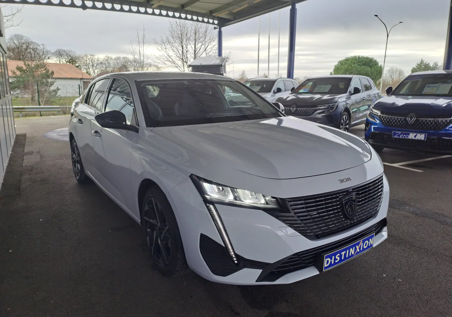 Peugeot 308 blanc Okenite vue 3/4 avant droit sous abri avec calandre noire et feux LED allumés.