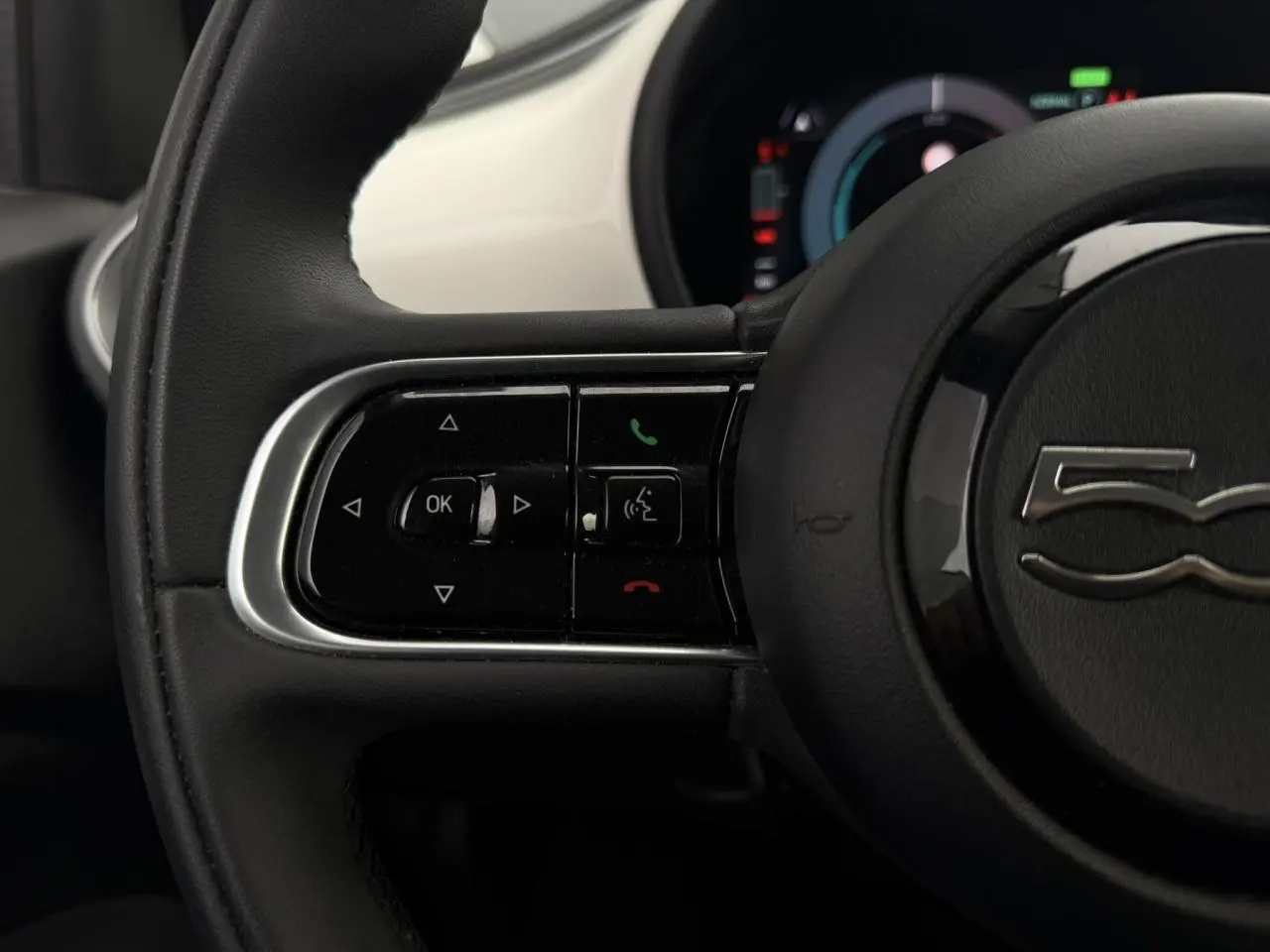 Gros plan sur la partie gauche du volant noir de la Fiat 500 électrique blanche, avec commandes intégrées et logo 500 visible.