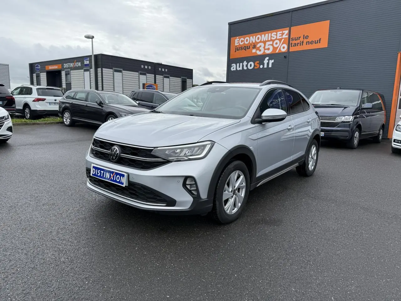 Volkswagen Taigo gris reflet d'argent métallisé en 3/4 avant droit sur parking devant un bâtiment commercial.