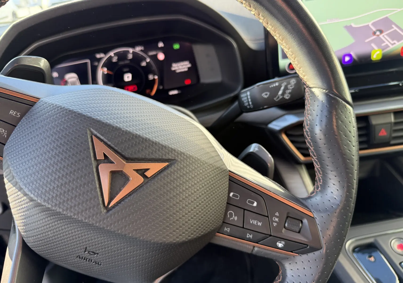 Gros plan sur le volant en cuir perforé noir avec logo CUPRA cuivré, tableau de bord digital et écran tactile visible en arrière-plan.