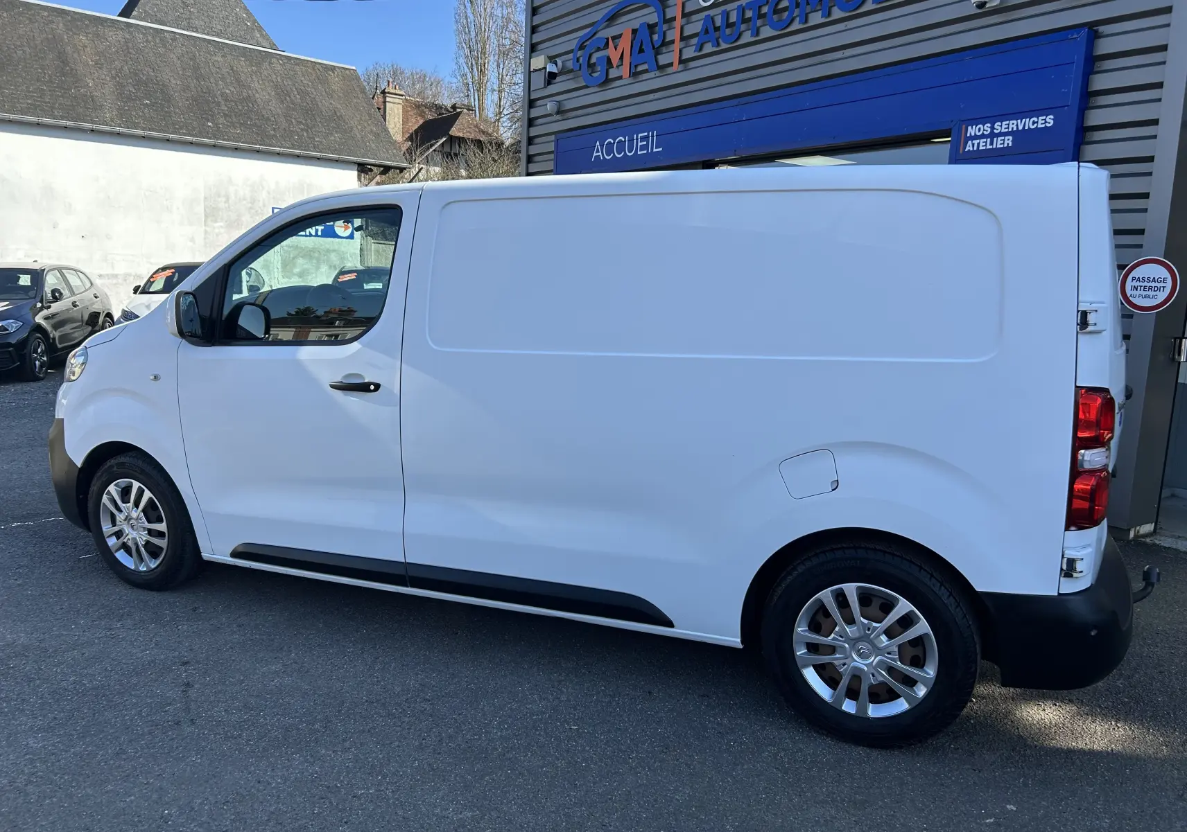 Vue de profil côté gauche d’un fourgon Citroën Jumpy blanc avec attelage, stationné devant un garage automobile.