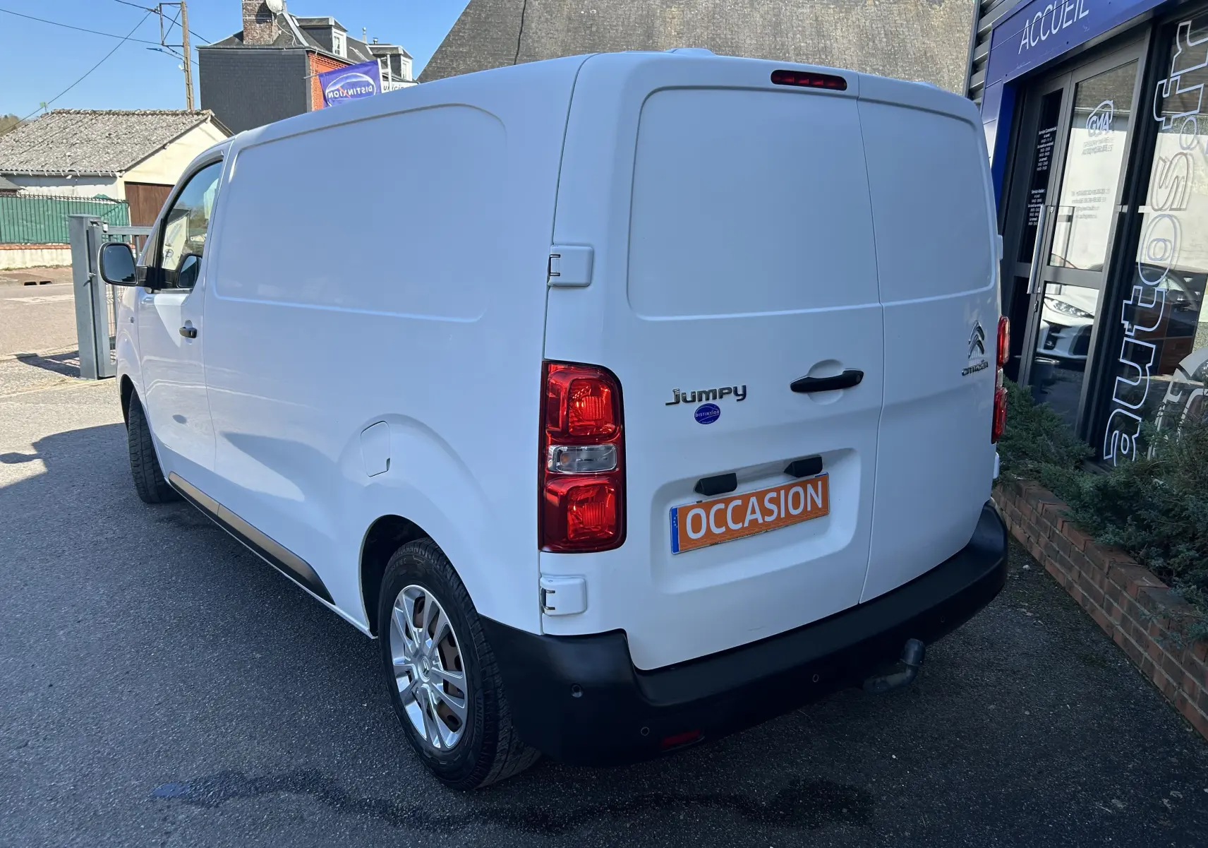 Vue 3/4 arrière droite d’un Citroën Jumpy blanc avec attelage, stationné devant un bâtiment commercial.