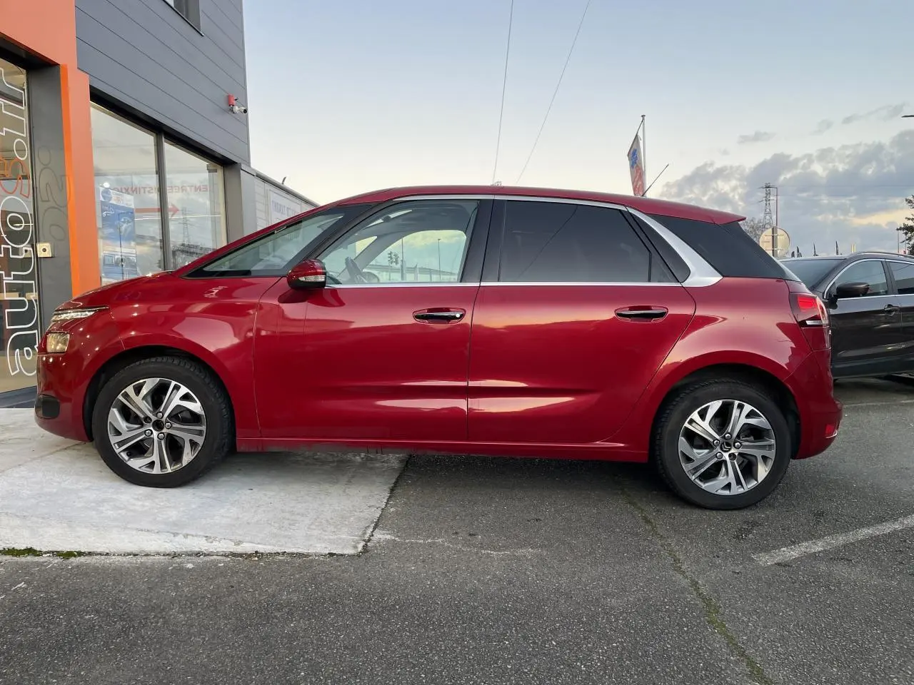 Profil côté gauche d'une Citroën C4 Picasso rouge foncé garée devant un bâtiment commercial.