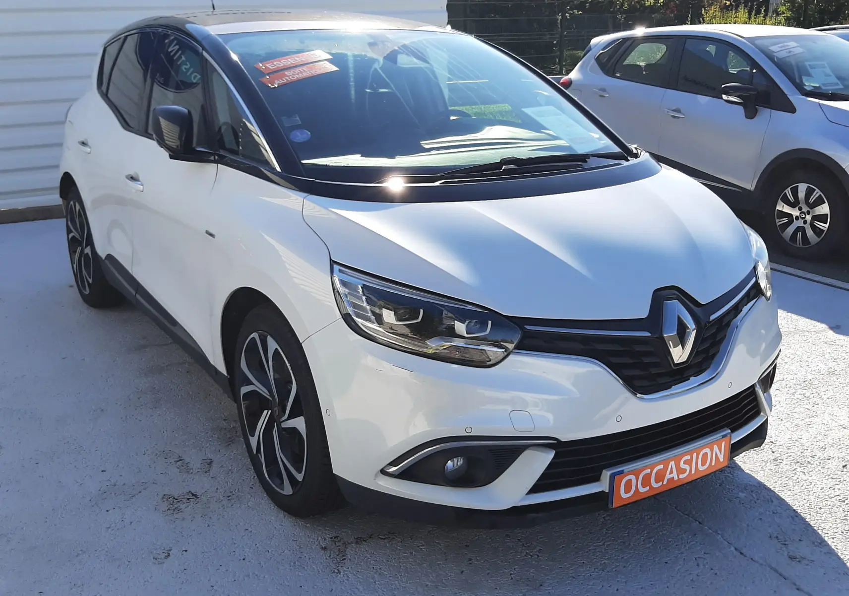 Renault Scénic IV blanc nacré avec toit noir, vue 3/4 avant droit, sur parking avec panneau multimarque en arrière-plan.