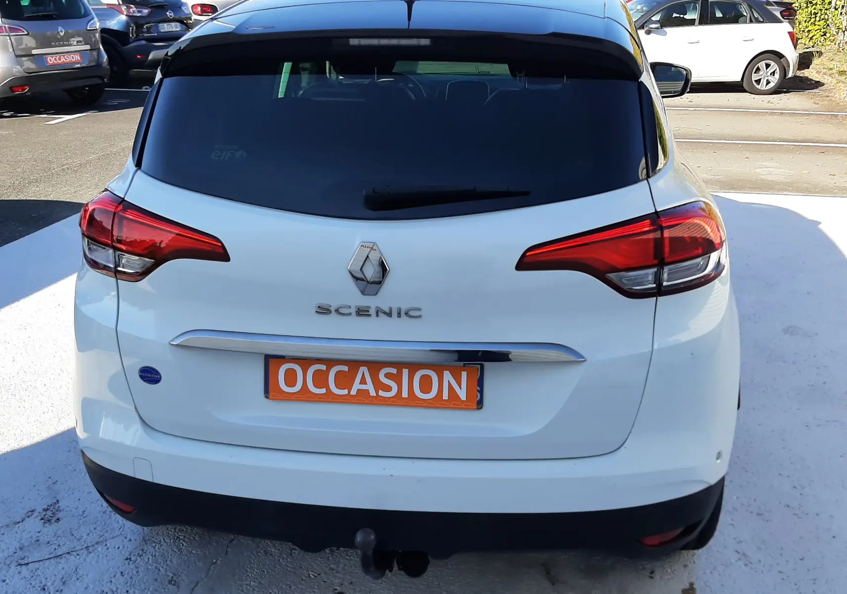 Vue arrière d'un Renault Scénic IV blanc nacré avec toit noir, feux arrière étirés et plaque "OCCASION" orange.