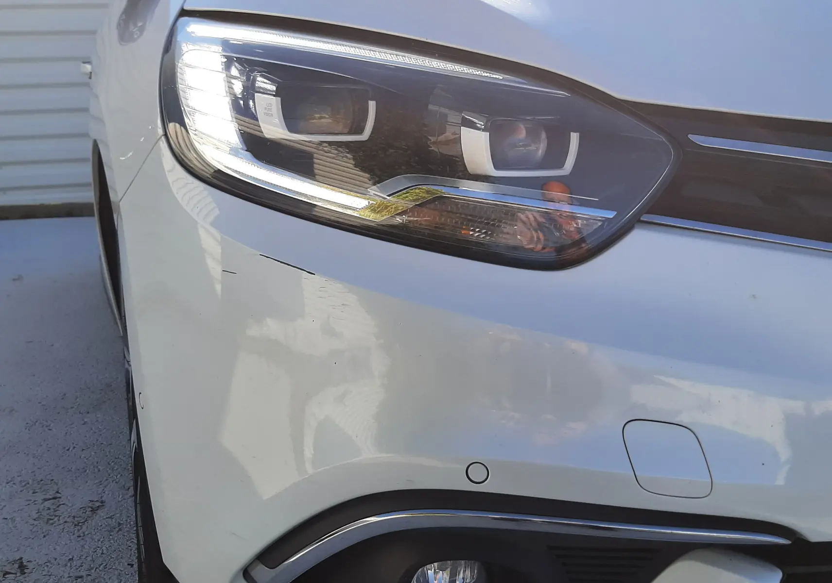 Gros plan sur le phare avant gauche d'un Renault Scénic IV blanc nacré avec toit noir, mettant en valeur ses feux LED et détails chromés.
