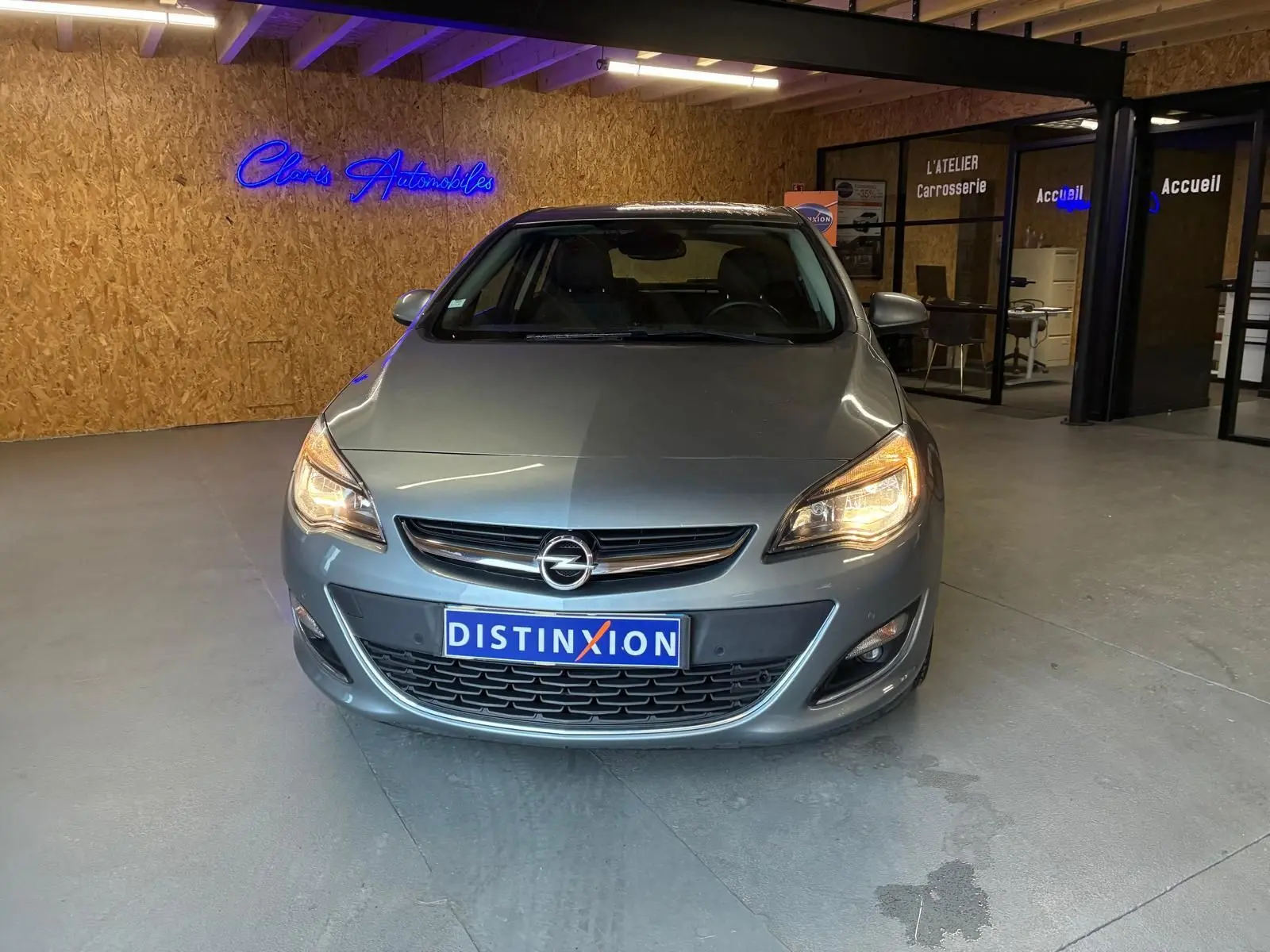 Vue avant d'une Opel Astra gris métallisé 2015 avec phares allumés dans un showroom intérieur.