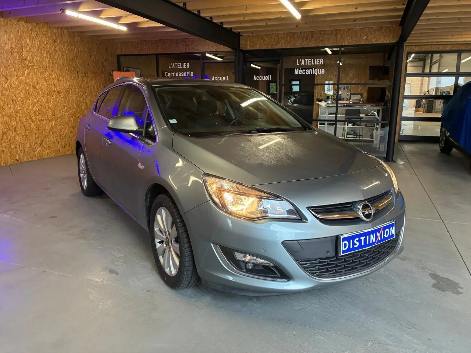 Opel Astra gris 2015 vue 3/4 avant droit avec phares allumés dans un atelier mécanique.