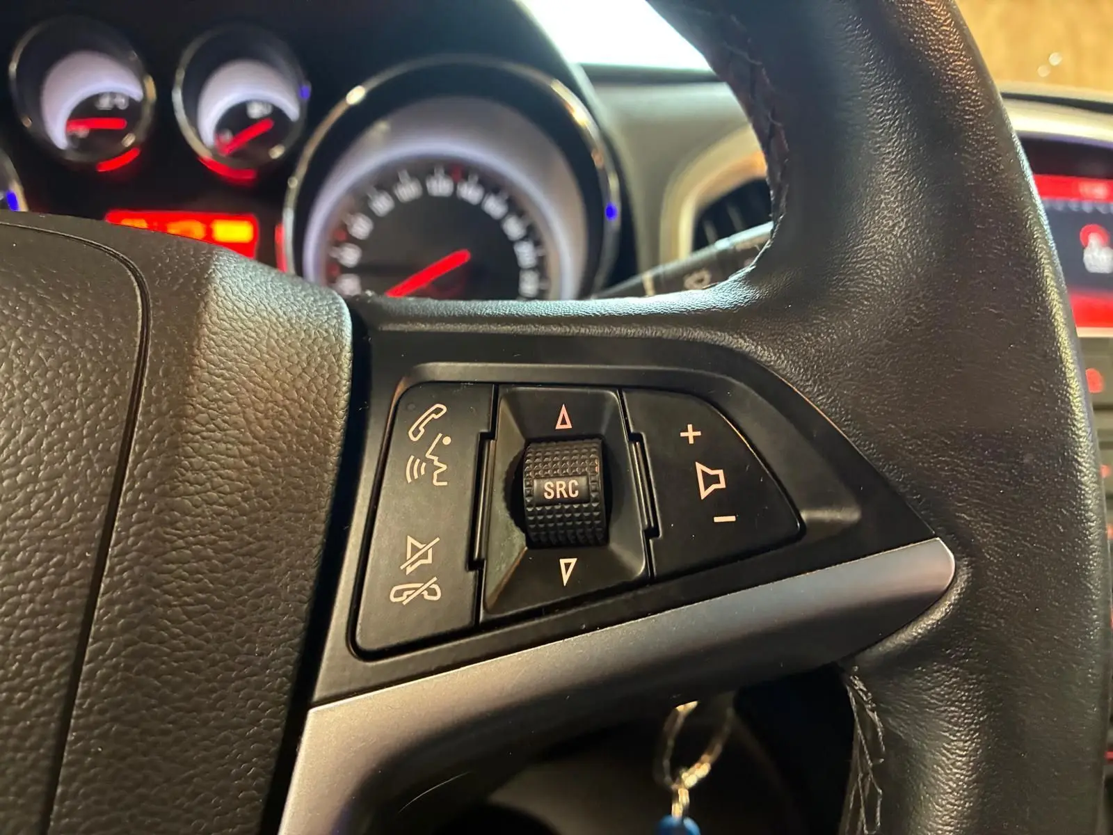 Gros plan sur les commandes multifonctions au volant d'une Opel Astra gris, avec tableau de bord et compteurs en arrière-plan flous.