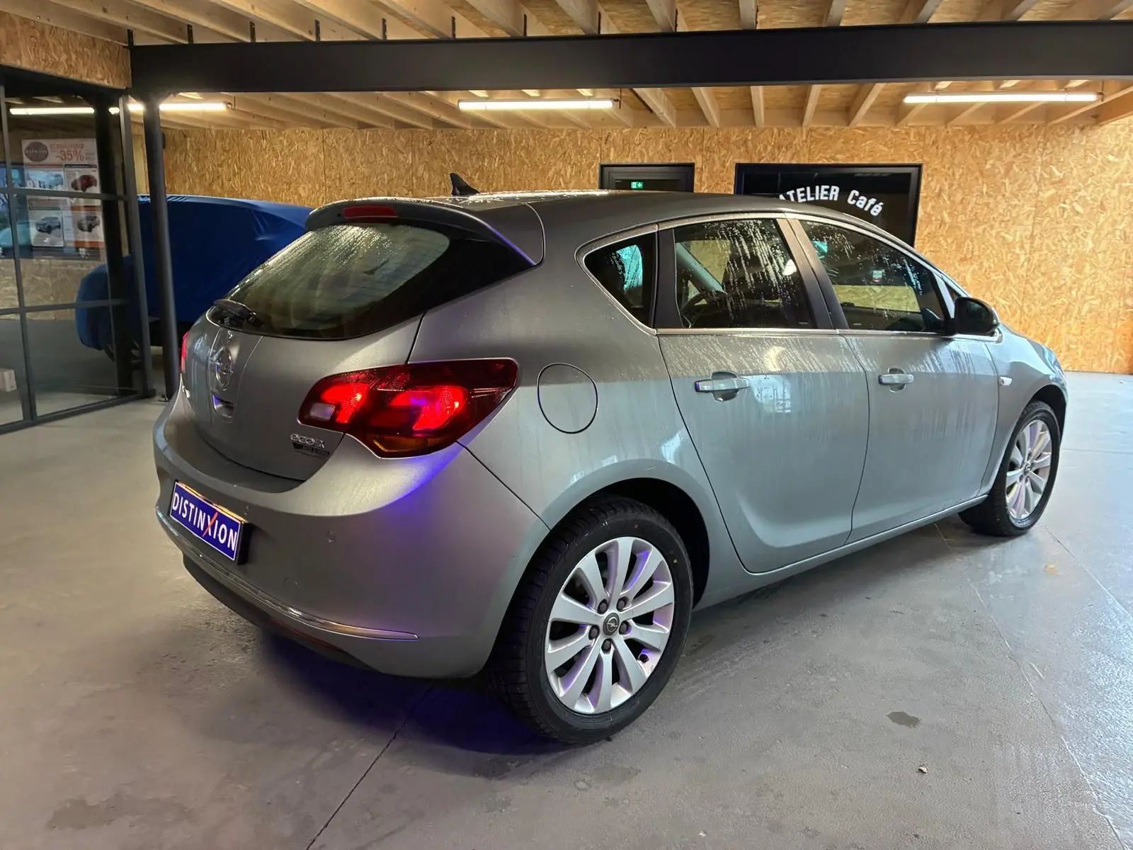 Opel Astra gris vue 3/4 arrière droit dans un garage avec feux arrière allumés et jantes alliage.