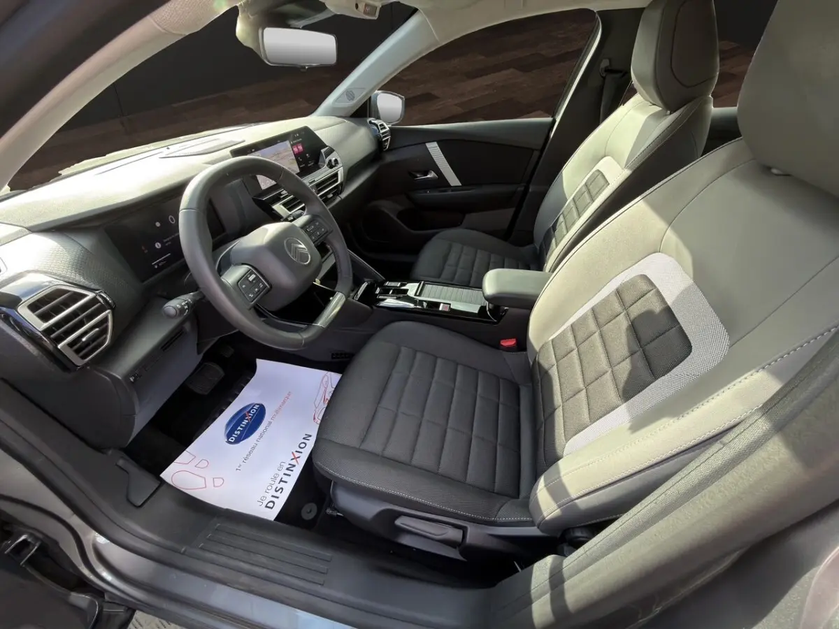 Vue intérieure côté conducteur de la Citroën C4 hybride 2025, sièges gris à motifs carrés et tableau de bord moderne.