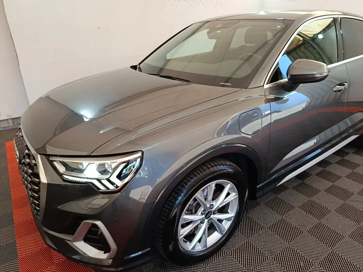 Audi Q3 Sportback gris clair en 3/4 avant droit, avec phares LED et jantes alliage visibles.