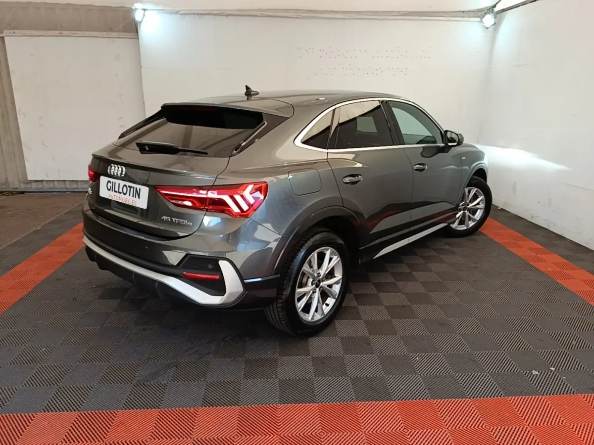 Audi Q3 Sportback gris clair en 3/4 arrière droit, avec feux LED et jantes alliage visibles en intérieur showroom.