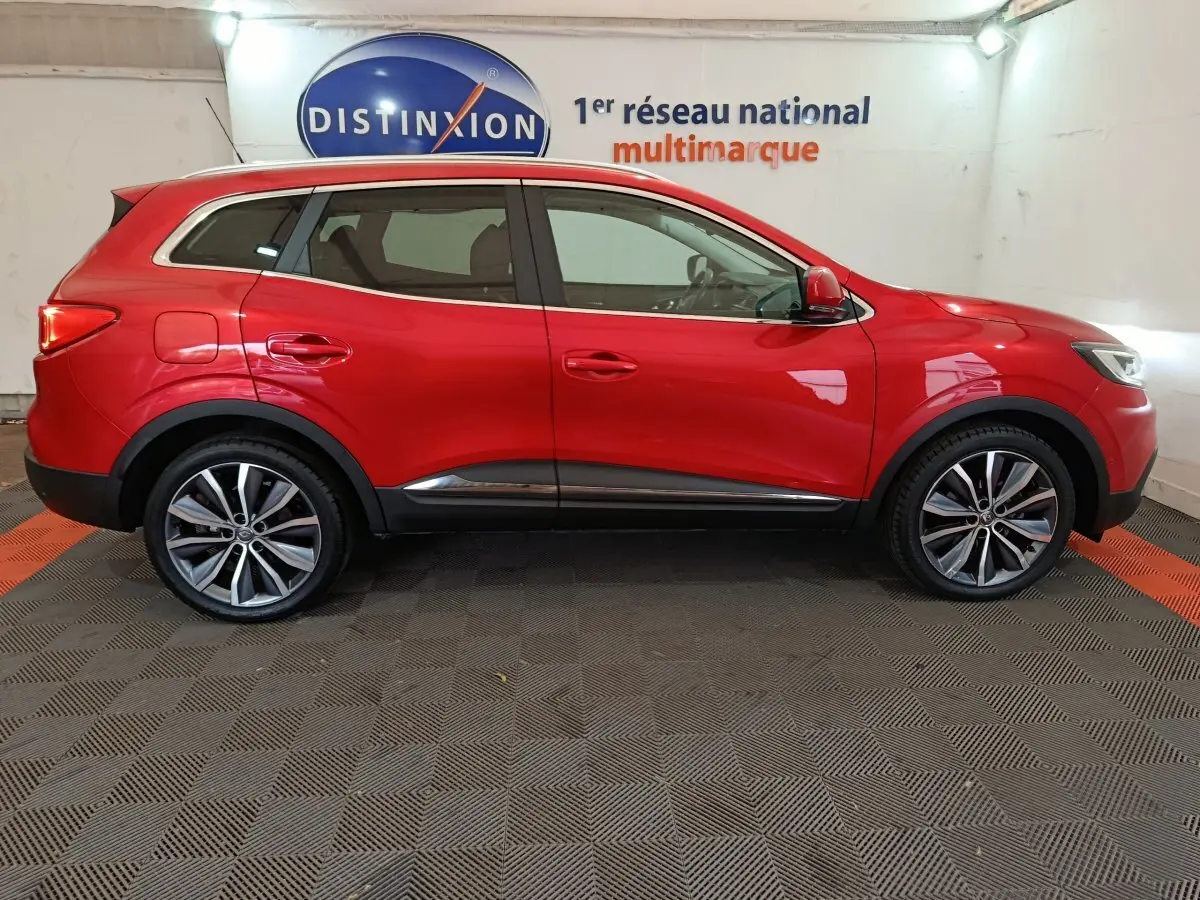 Vue latérale droite d'un Renault Kadjar Intens rouge brillant, avec jantes alliage et toit chromé en arrière-plan showroom.