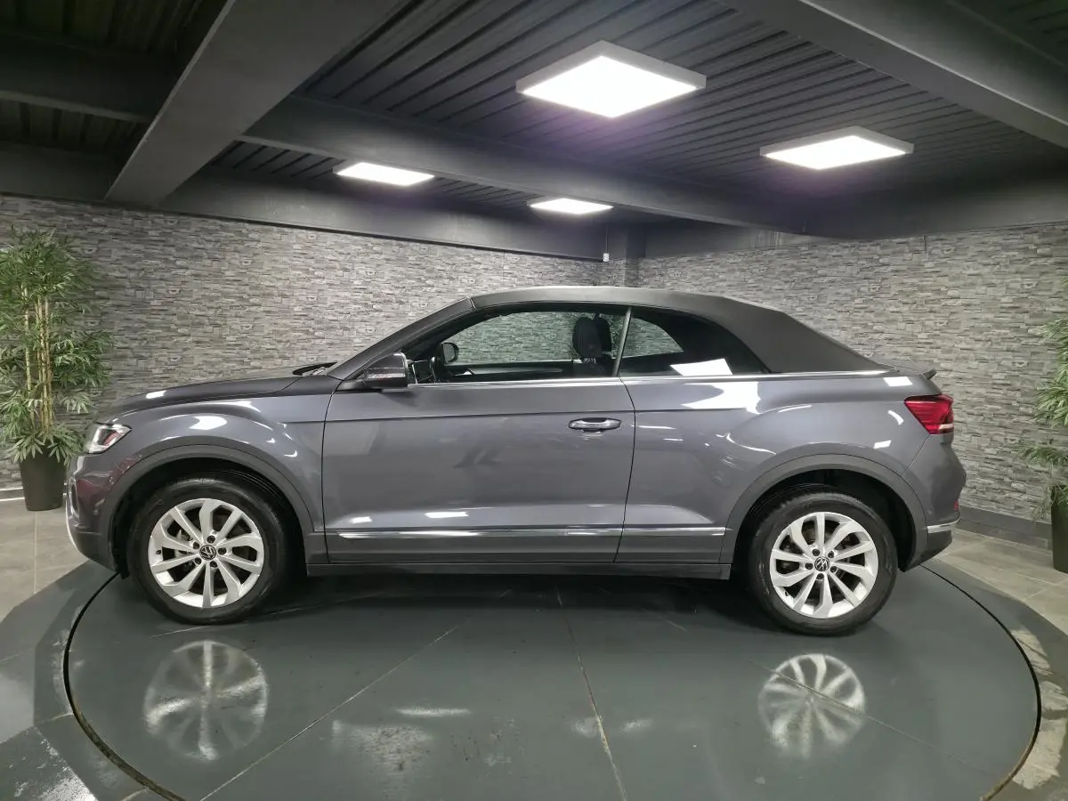 Volkswagen T-Roc Cabriolet gris métallisé vue de profil côté gauche avec capote noire fermée en intérieur showroom.