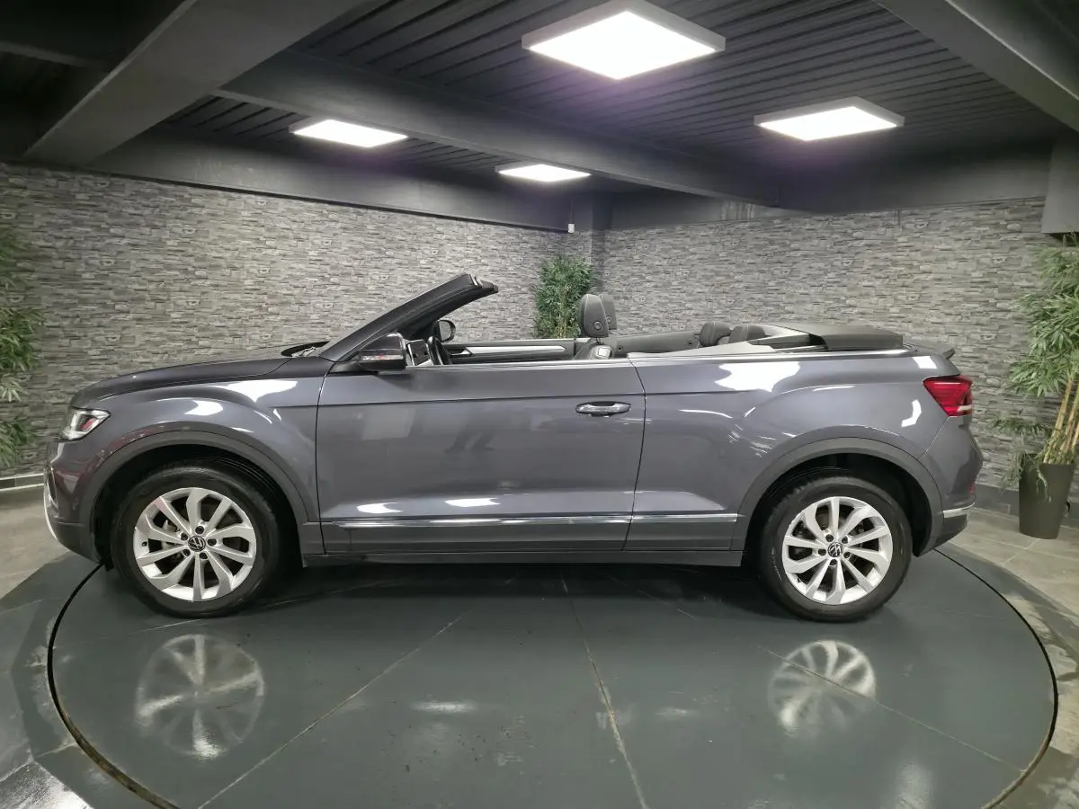 Profil côté gauche d'un Volkswagen T-Roc Cabriolet gris métallisé 2024 avec capote ouverte en intérieur showroom.