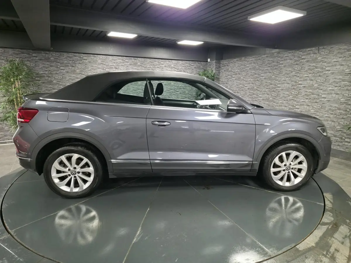 Profil côté gauche d'un Volkswagen T-Roc Cabriolet gris métallisé avec capote noire dans un showroom.