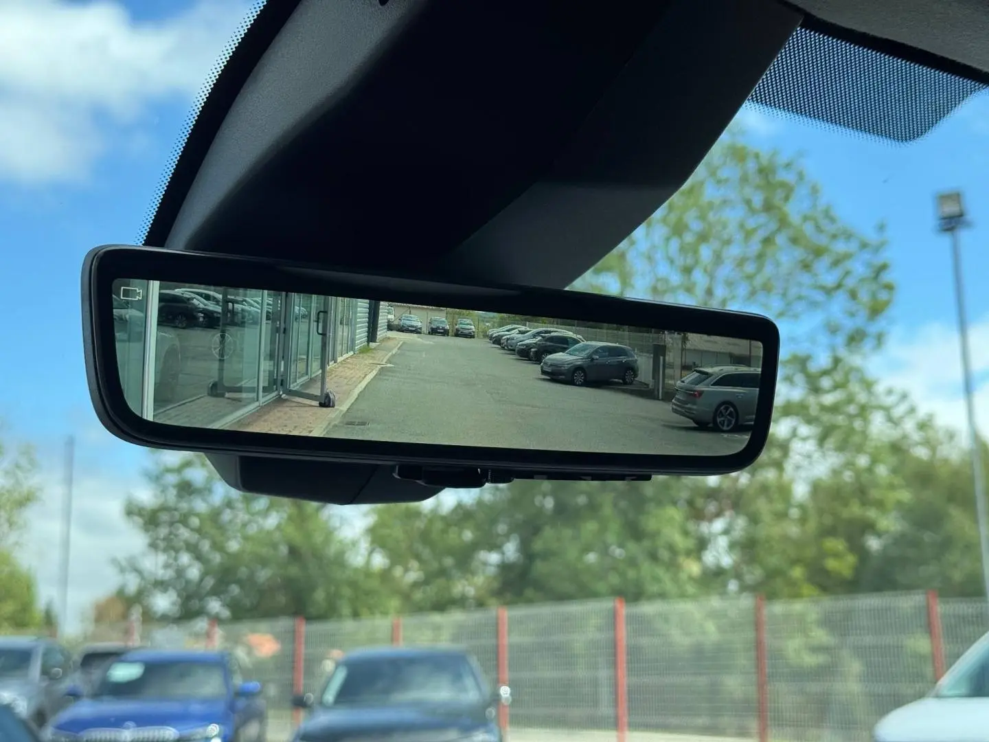 Rétroviseur intérieur numérique montrant une vue arrière dégagée d’un parking sous ciel bleu.