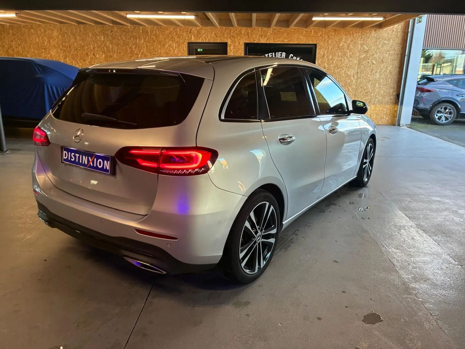 Mercedes Classe B 200D Progressive Line gris vue 3/4 arrière droit, feux arrière allumés en intérieur d'atelier.