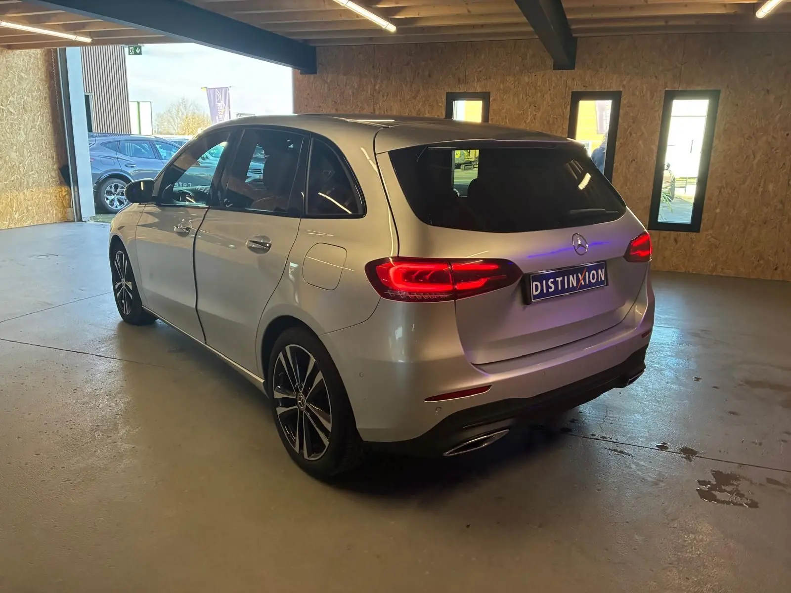 Vue 3/4 arrière droite d'une Mercedes Classe B 200D Progressive Line gris métallisé, feux arrière allumés, en intérieur.