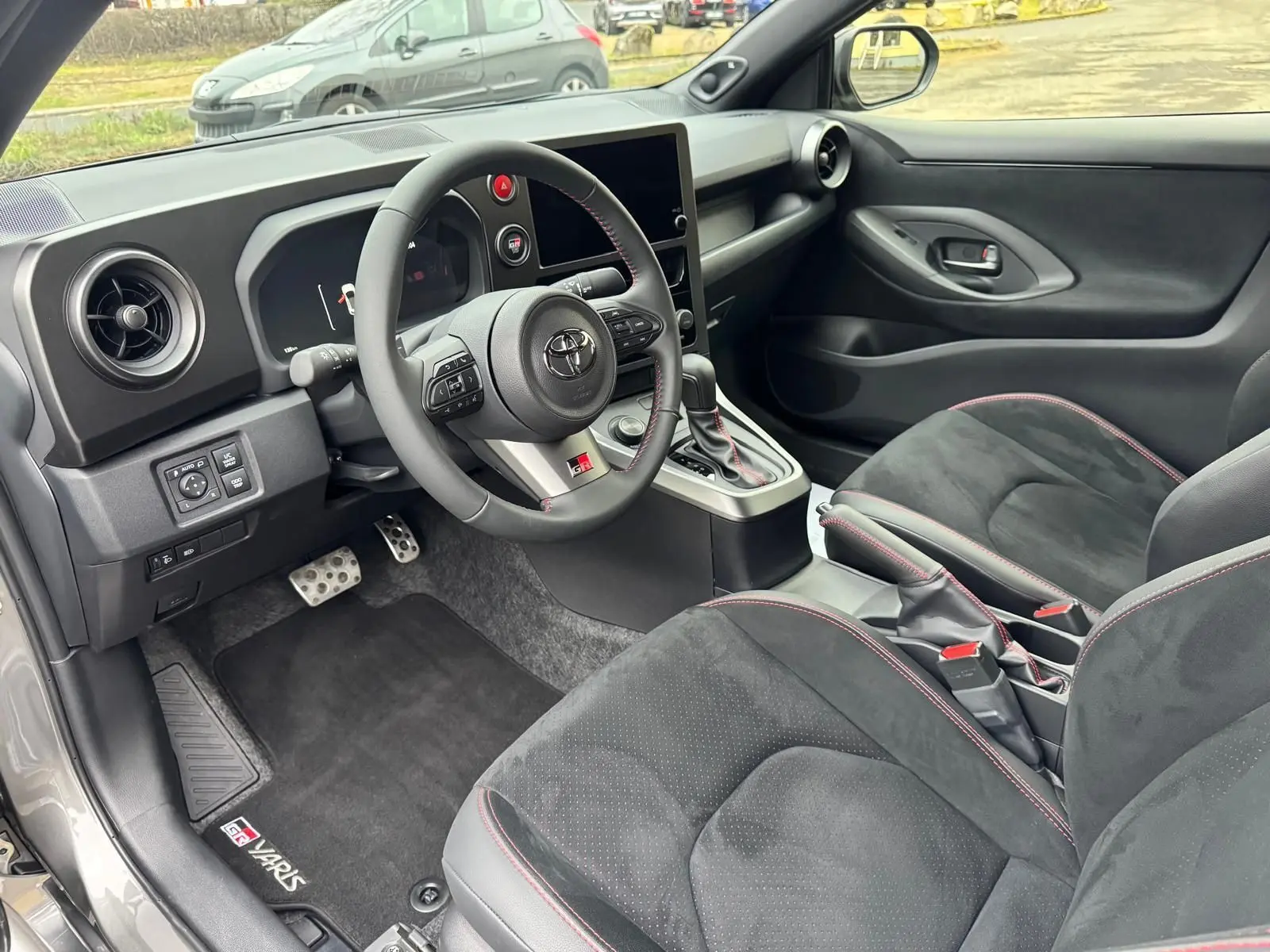 Vue intérieure côté conducteur de la Toyota Yaris GR avec sièges noirs surpiqués rouges et volant sport à coutures rouges.