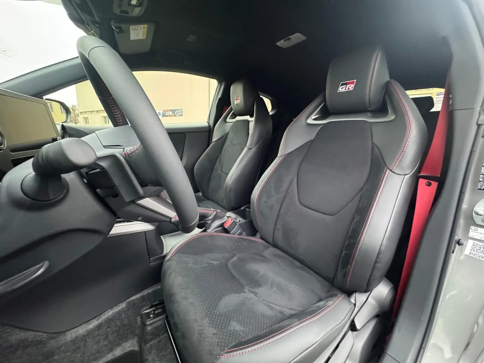 Vue intérieure côté conducteur de la Toyota Yaris GR avec sièges sport noirs, surpiqûres rouges et ceinture rouge visible.