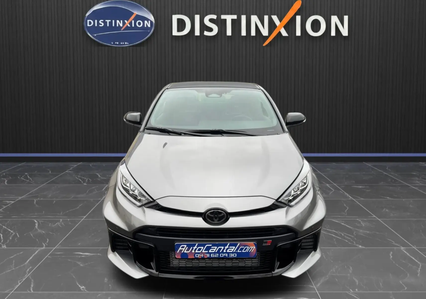 Vue avant d'une Toyota Yaris GR gris platine avec calandre noire et logo GR, sur fond noir avec logo Distinxion.