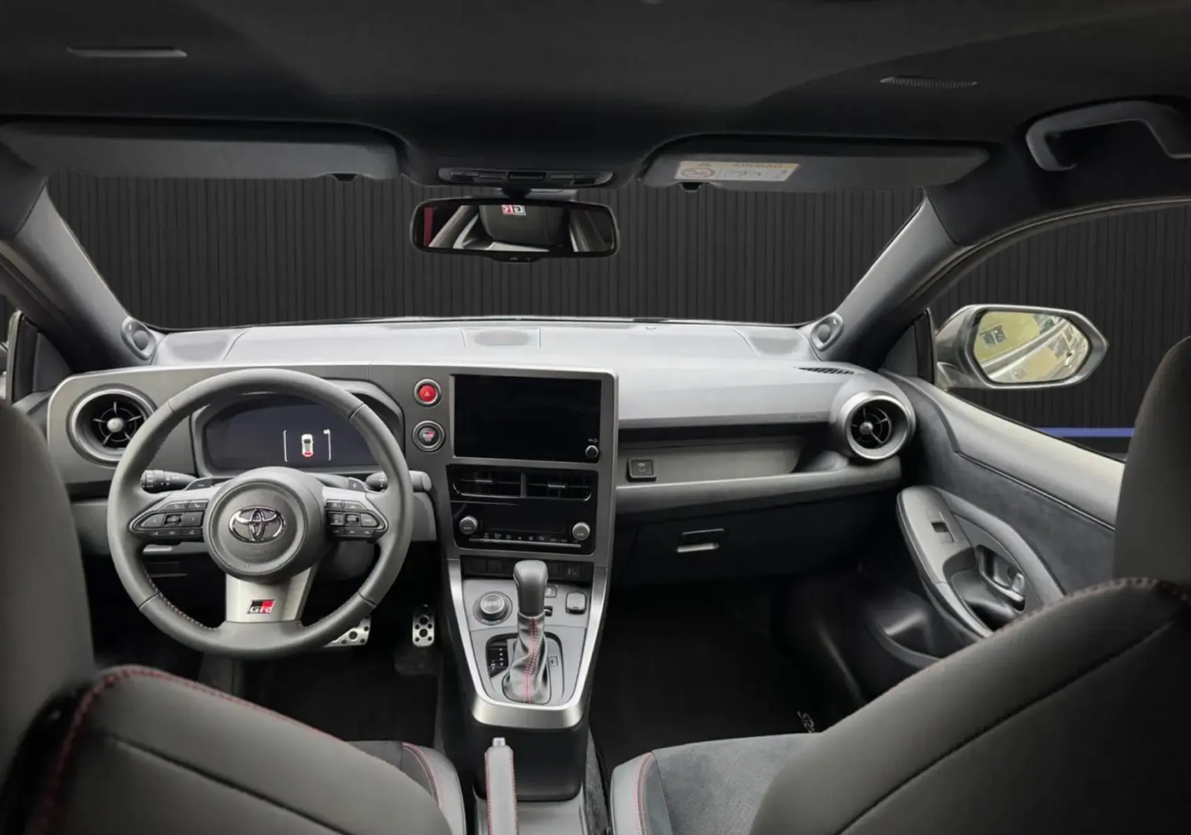 Vue intérieure avant de la Toyota Yaris GR Pack Techno, tableau de bord noir avec surpiqûres rouges et volant sport multifonction.