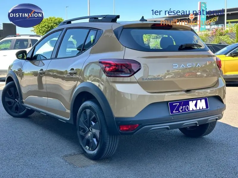 Vue 3/4 arrière droite d'une Dacia Sandero beige Safari avec barres de toit et jantes noires sur parking.