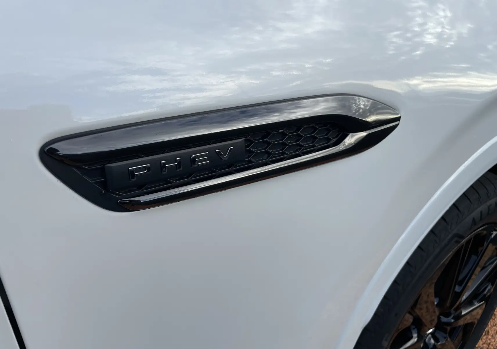 Gros plan sur l'aile avant droite blanche nacrée du Mazda CX-60 avec l'insigne noir PHEV et une partie de la roue.