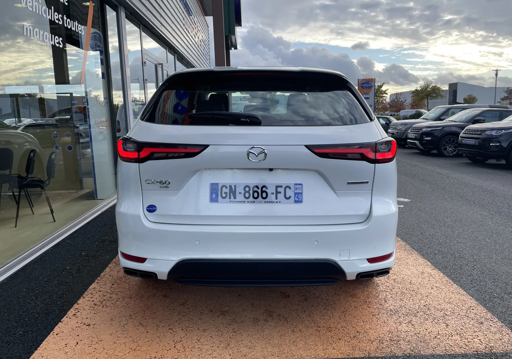 Vue arrière d'un Mazda CX-60 blanc nacré 2023 avec feux LED et double sortie d'échappement, stationné devant un showroom.