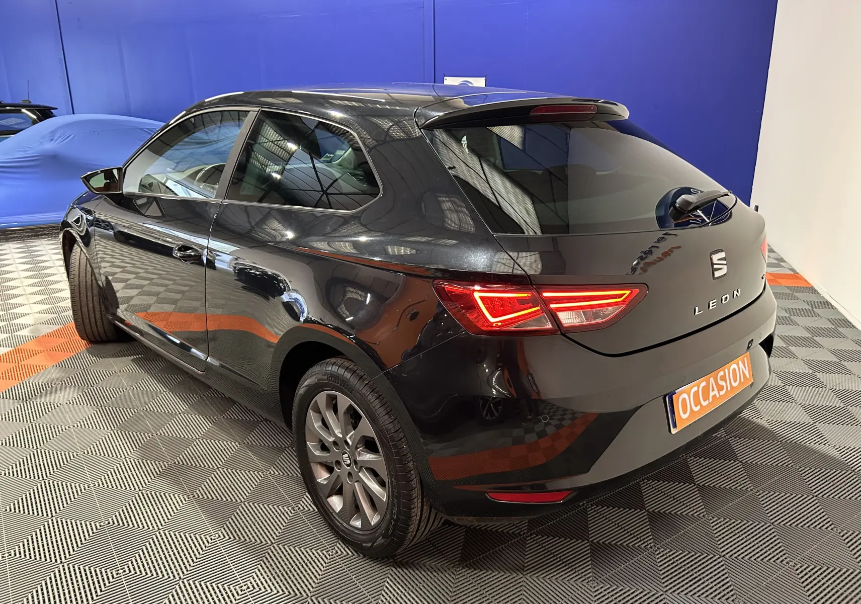 Vue 3/4 arrière droite d'une SEAT Leon noire de 2014 avec feux arrière allumés et plaque "OCCASION" visible.