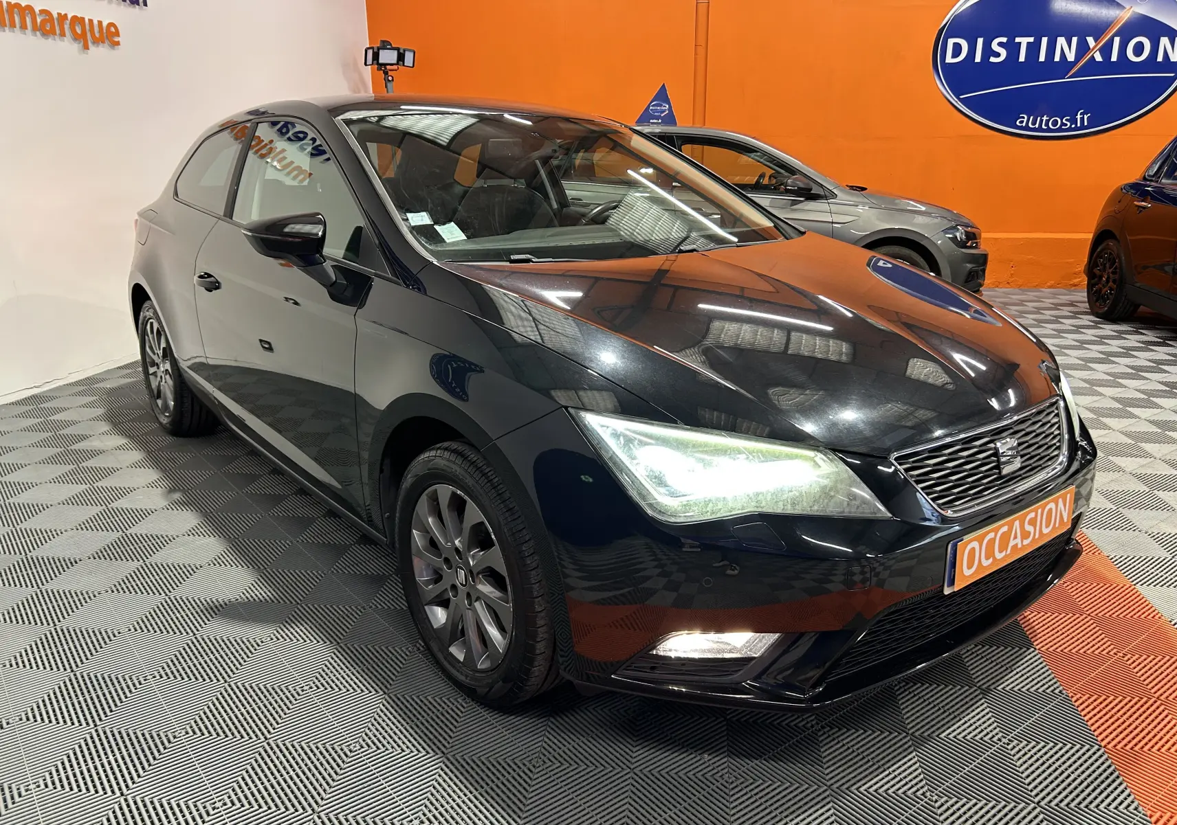 SEAT Leon noir vue 3/4 avant droit, phares allumés, sur sol damier dans showroom avec panneau orange "Occasion".