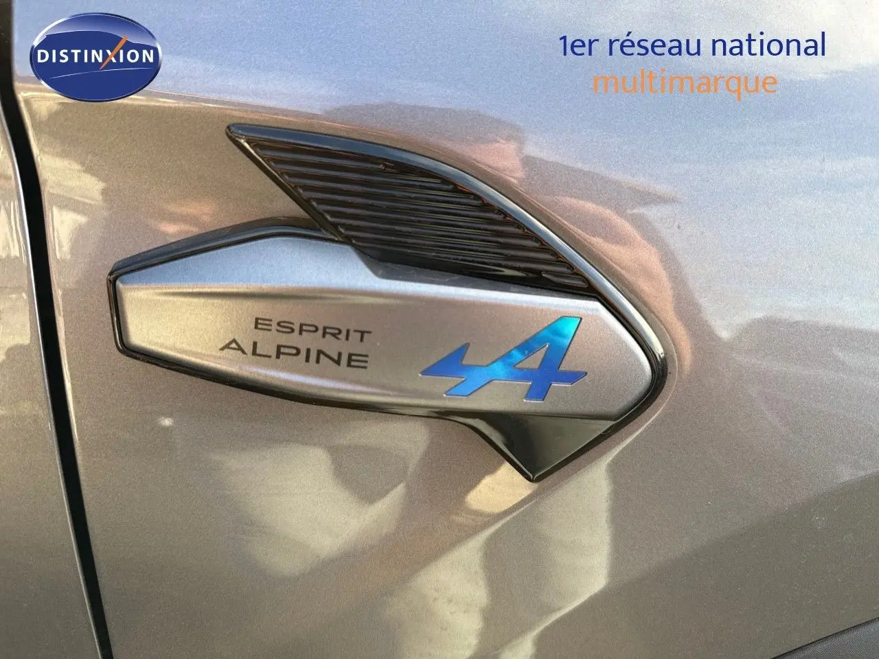 Gros plan sur l'emblème Esprit Alpine bleu sur la carrosserie gris cassiopée métal d'une Renault Symbioz 2024.