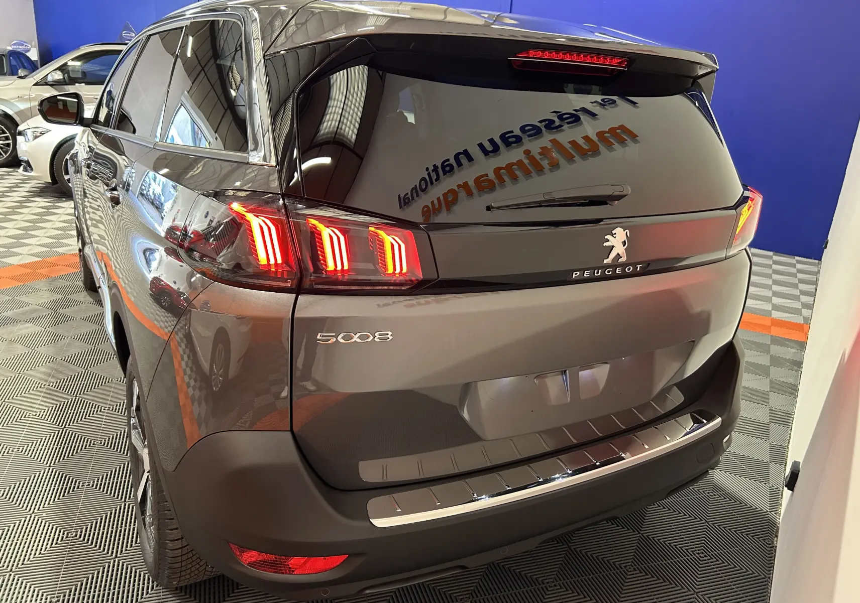 Vue 3/4 arrière droite du Peugeot 5008 gris Platinium avec feux arrière LED allumés en intérieur showroom.