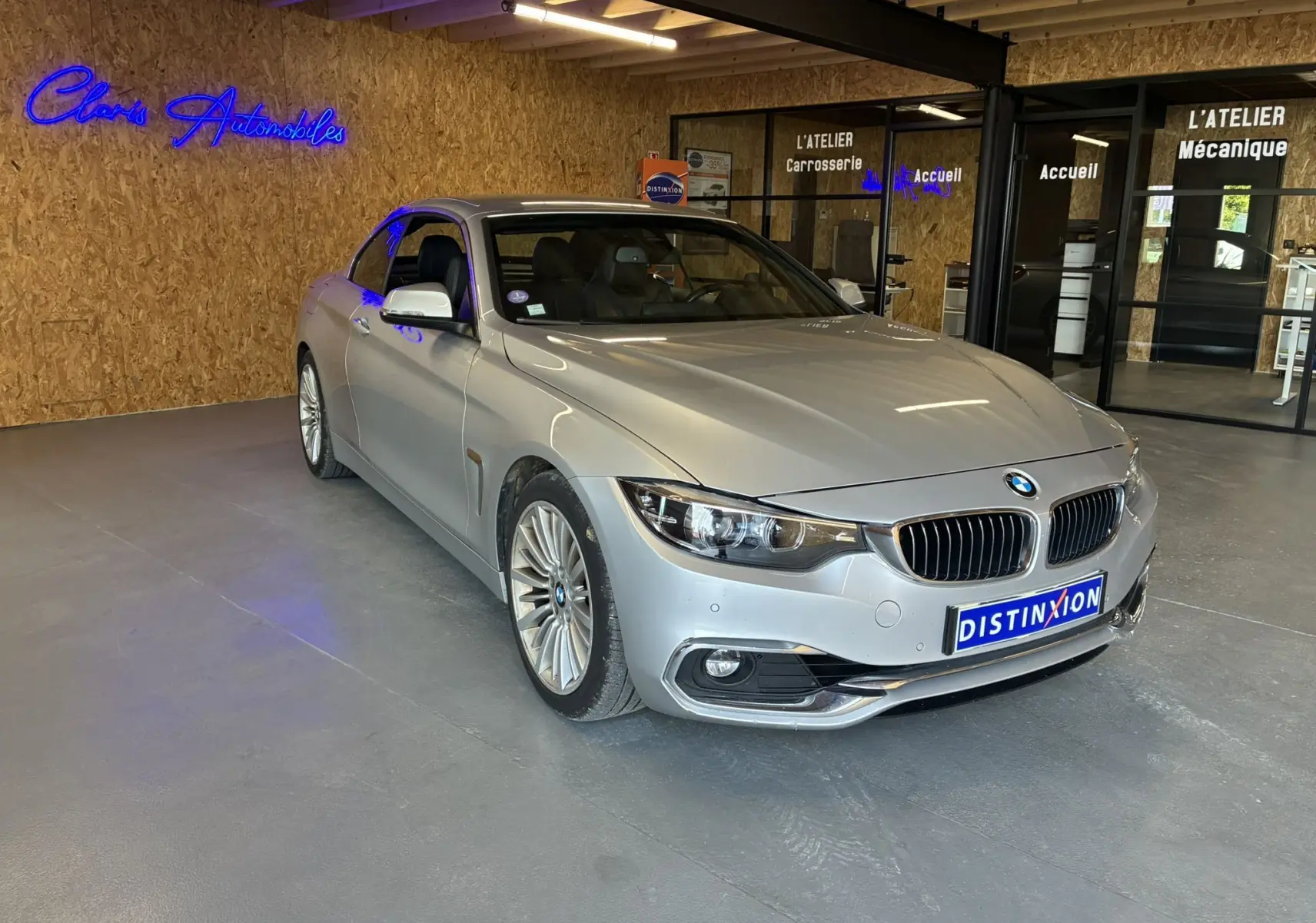 BMW Série 4 Cabriolet gris vue 3/4 avant droit dans un garage avec enseignes lumineuses en arrière-plan