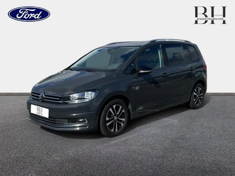 Volkswagen Touran gris Urano en 3/4 avant droit, avec jantes alliage et barres de toit visibles.