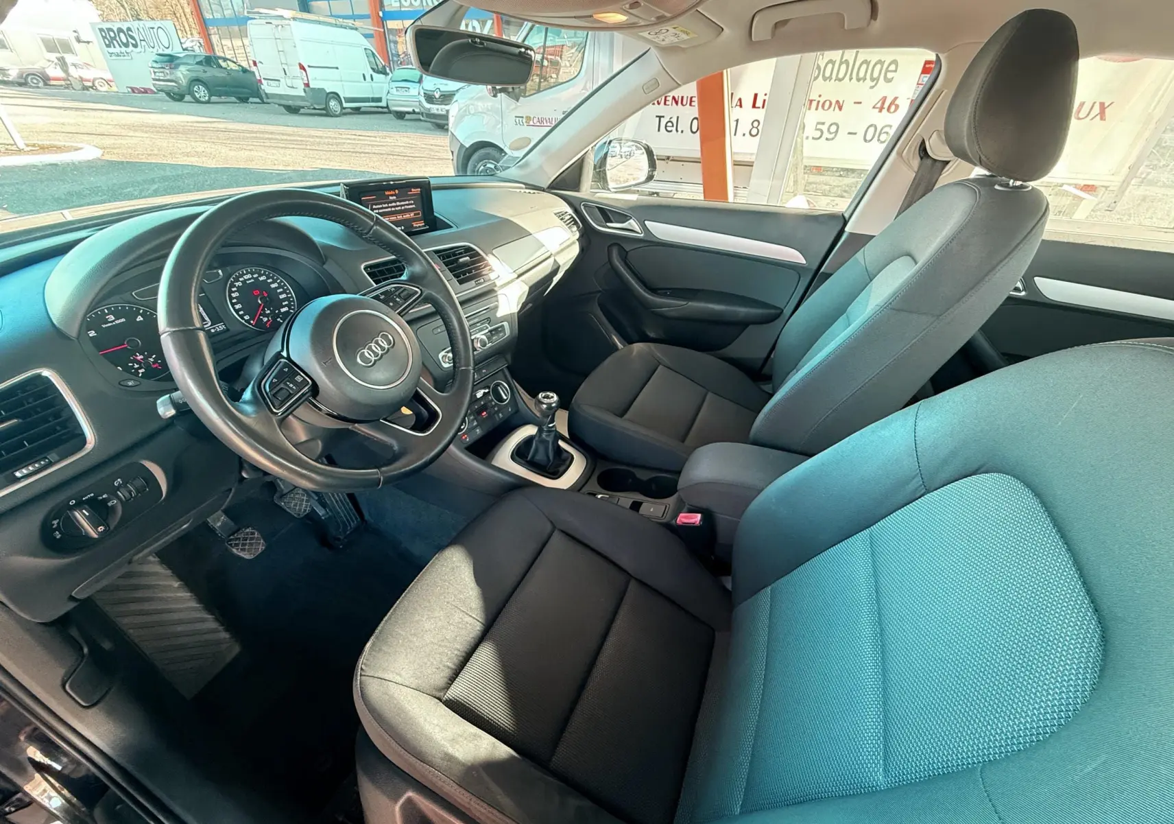 Vue intérieure côté conducteur d’un Audi Q3 noir, montrant le volant, la boîte manuelle et les sièges en tissu noir.