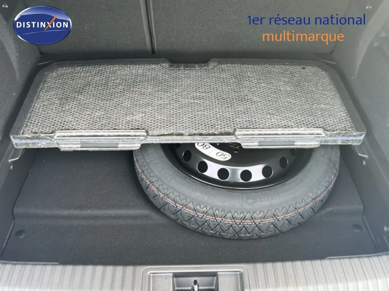 Coffre ouvert montrant la roue de secours sous un tapis partiellement relevé, intérieur noir du Renault Symbioz blanc nacré.