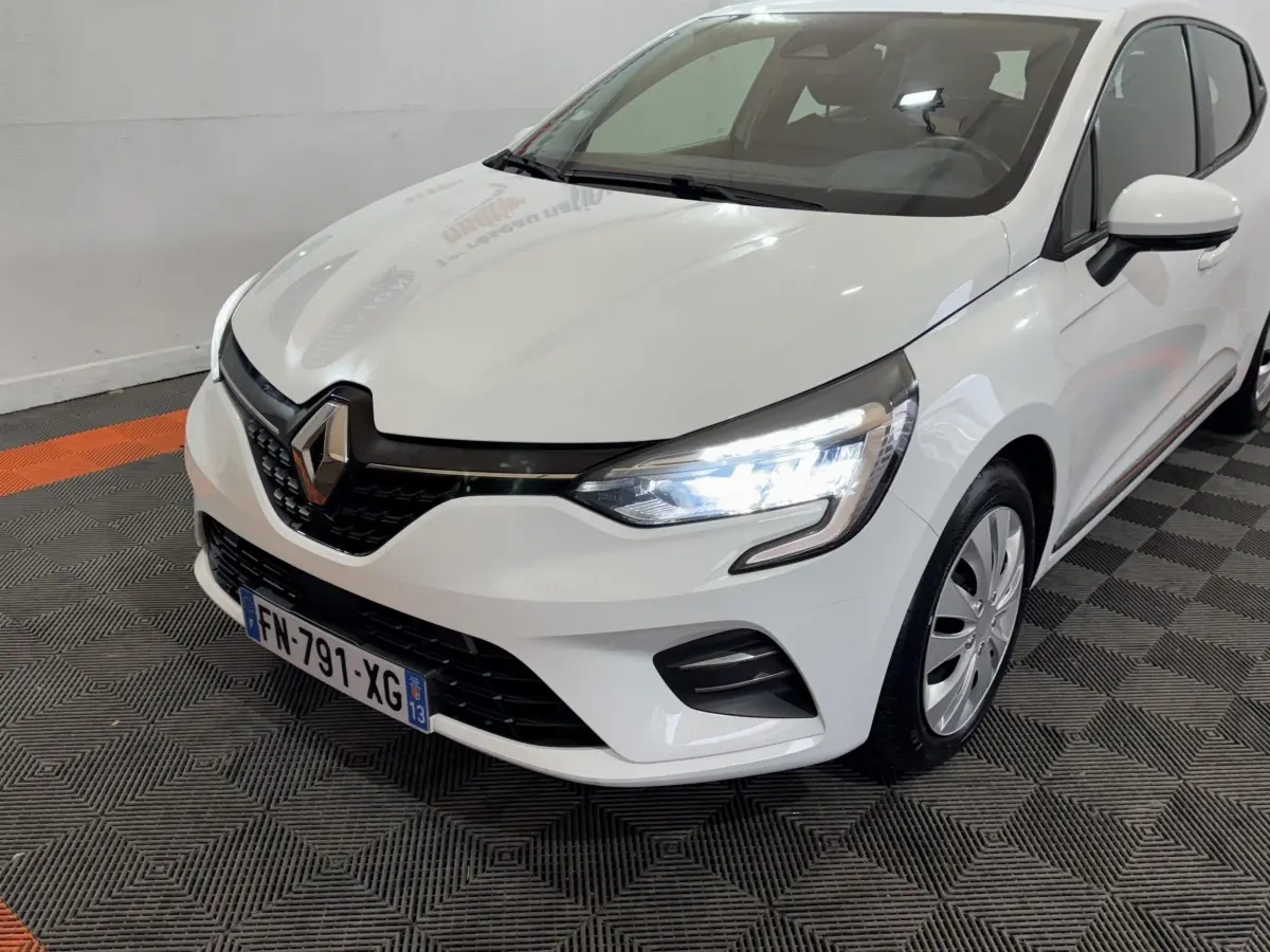 Renault Clio Business TCe 100 blanche vue 3/4 avant droit avec phares LED allumés dans un showroom.
