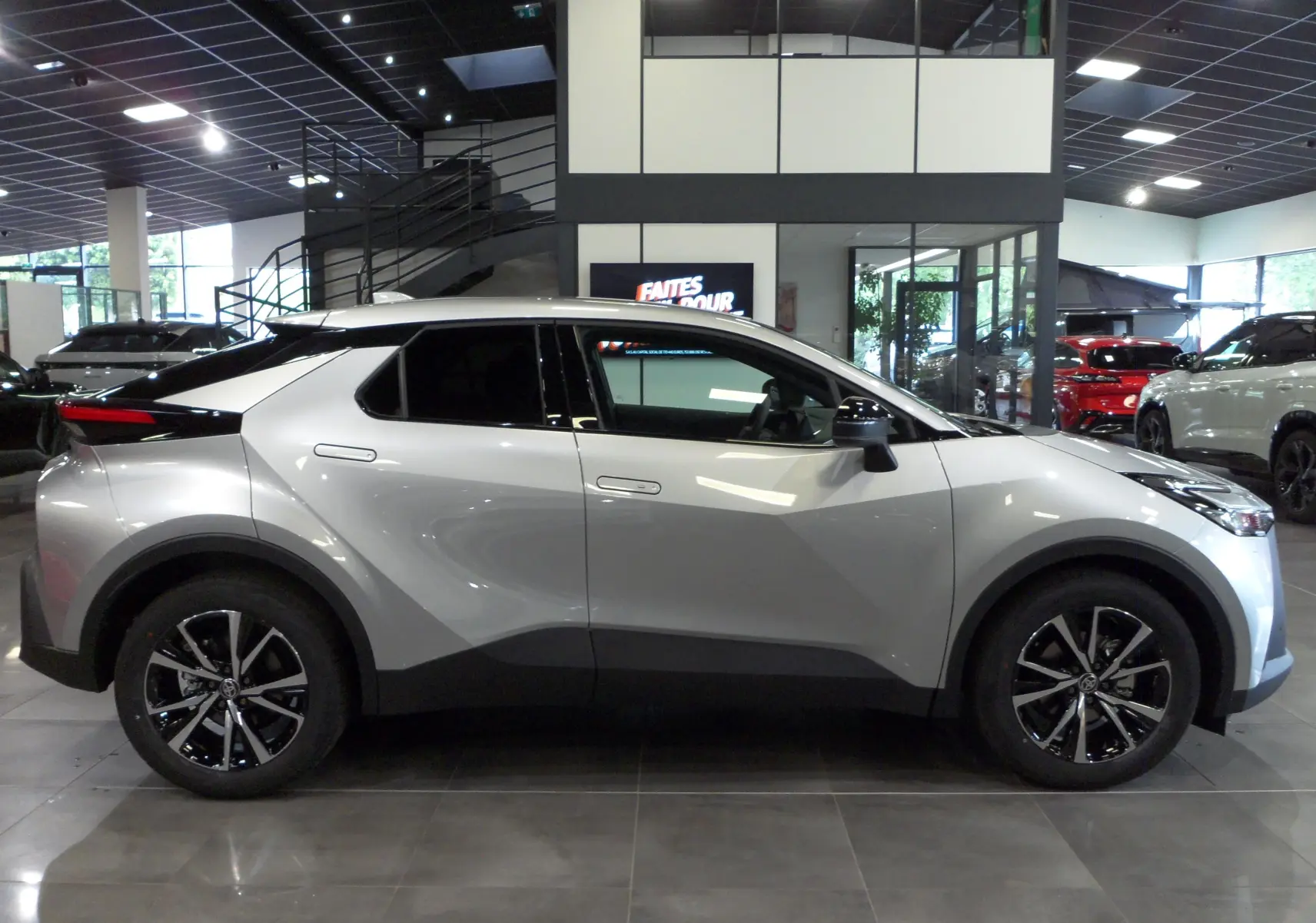 Profil côté droit du Toyota C-HR hybride 140 ch style 2025 en gris minéral avec jantes bi-ton 18 pouces en showroom.