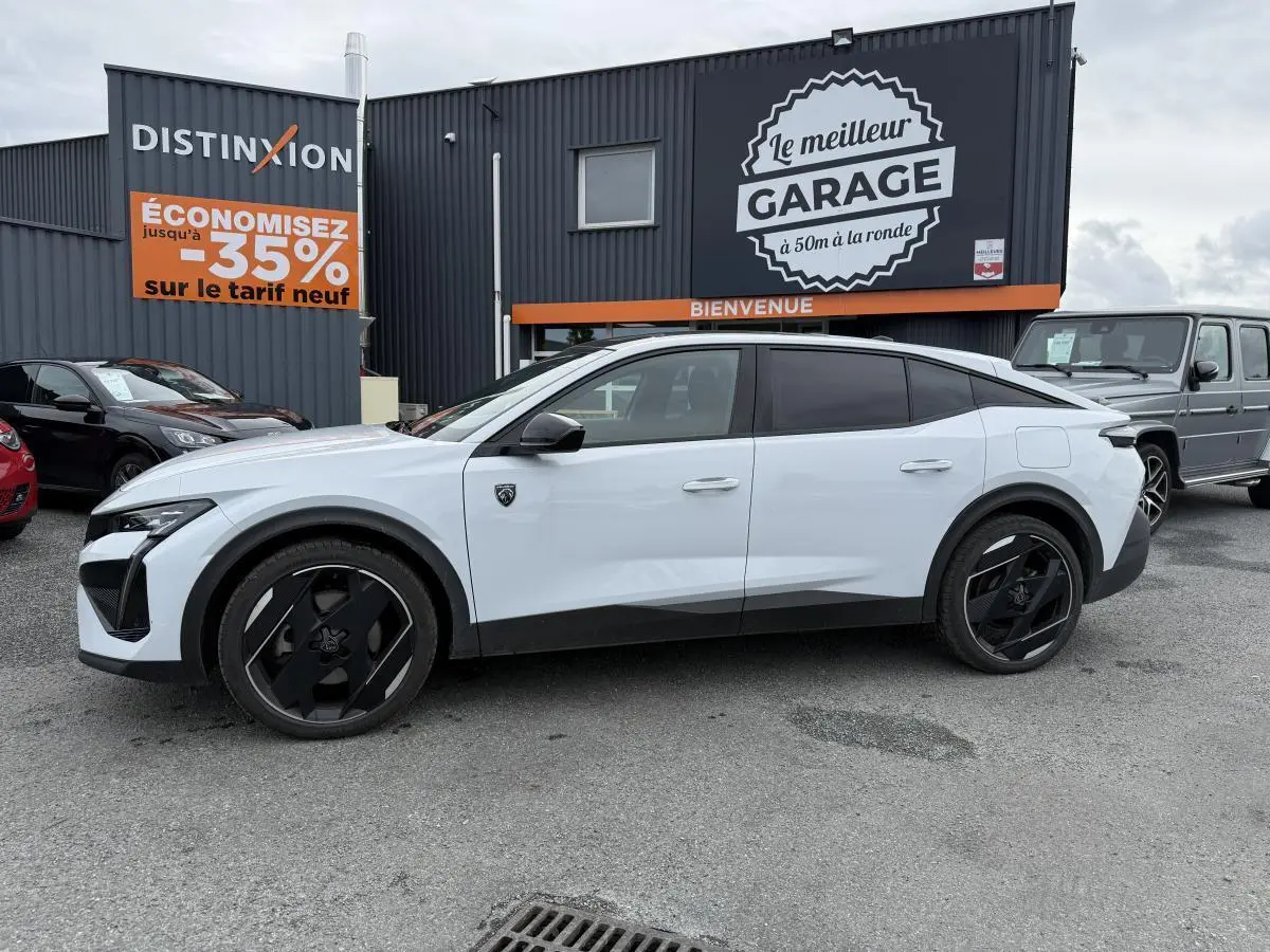 Vue de profil côté gauche d'une Peugeot 408 blanche avec jantes noires et détails noirs brillants sur un parking devant un garage.