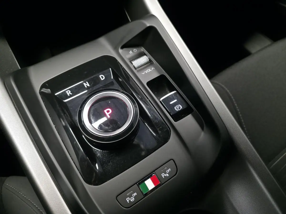 Gros plan sur la console centrale noire de l'Alfa Romeo Tonale bleu, avec sélecteur de vitesse rotatif et drapeau italien.