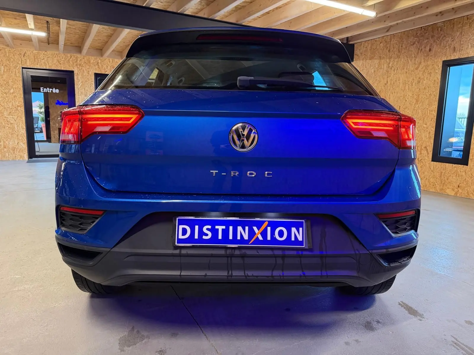 Vue arrière d'un Volkswagen T-Roc bleu 2019 avec feux arrière allumés et plaque Distinxion visible en intérieur.
