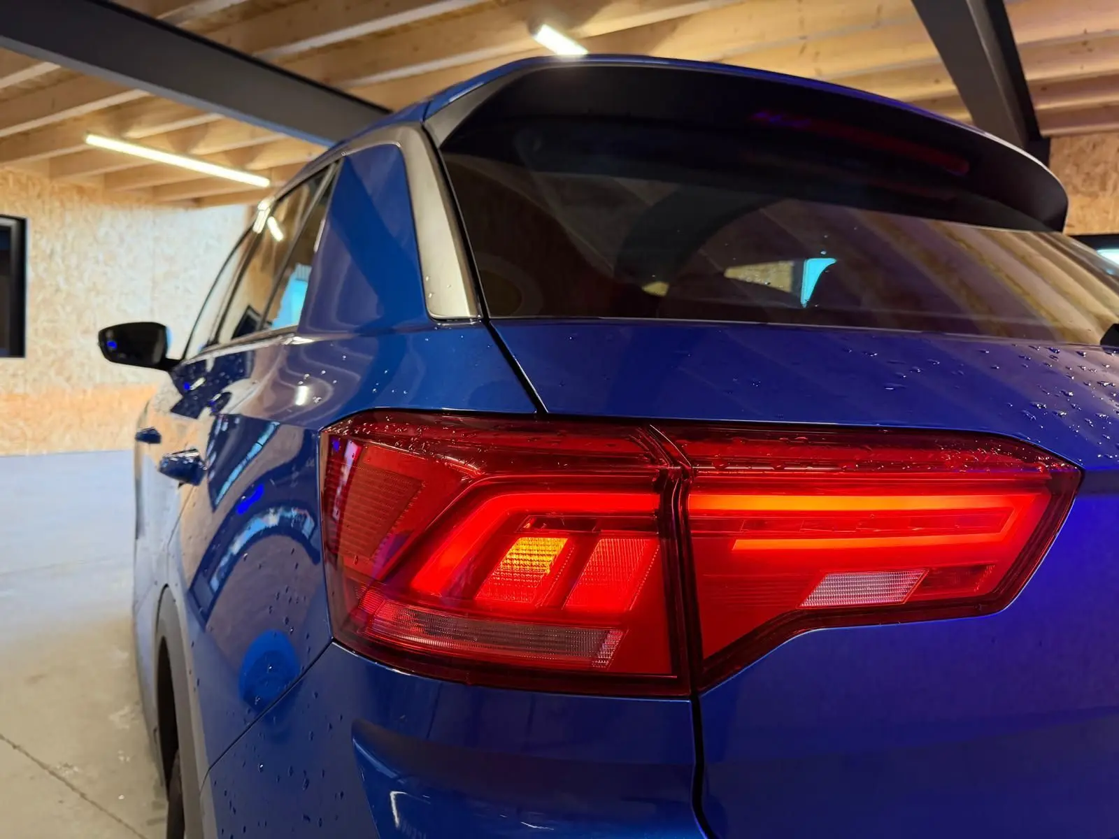 Vue 3/4 arrière gauche du Volkswagen T-Roc bleu avec feux arrière LED allumés dans un garage.