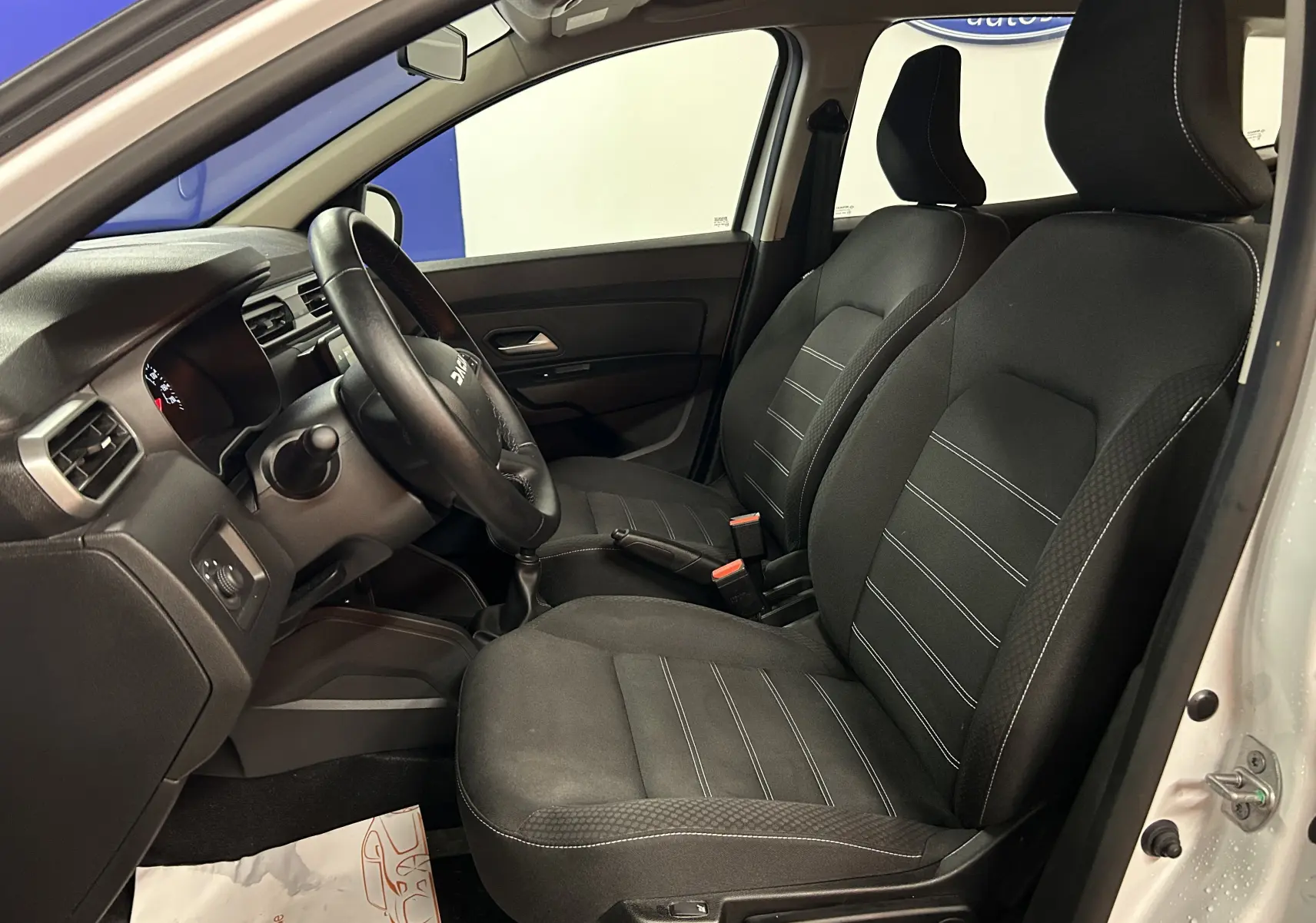 Vue intérieure côté conducteur du Dacia Duster blanc 2023, mettant en valeur les sièges chauffants en tissu noir avec surpiqûres blanches.