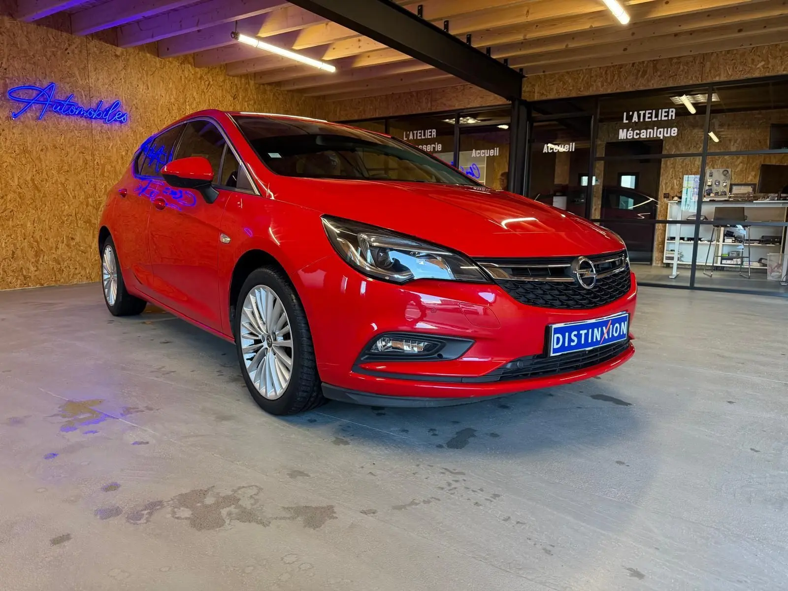 Opel Astra rouge vue 3/4 avant droit dans un garage avec éclairage intérieur et enseigne néon bleue.